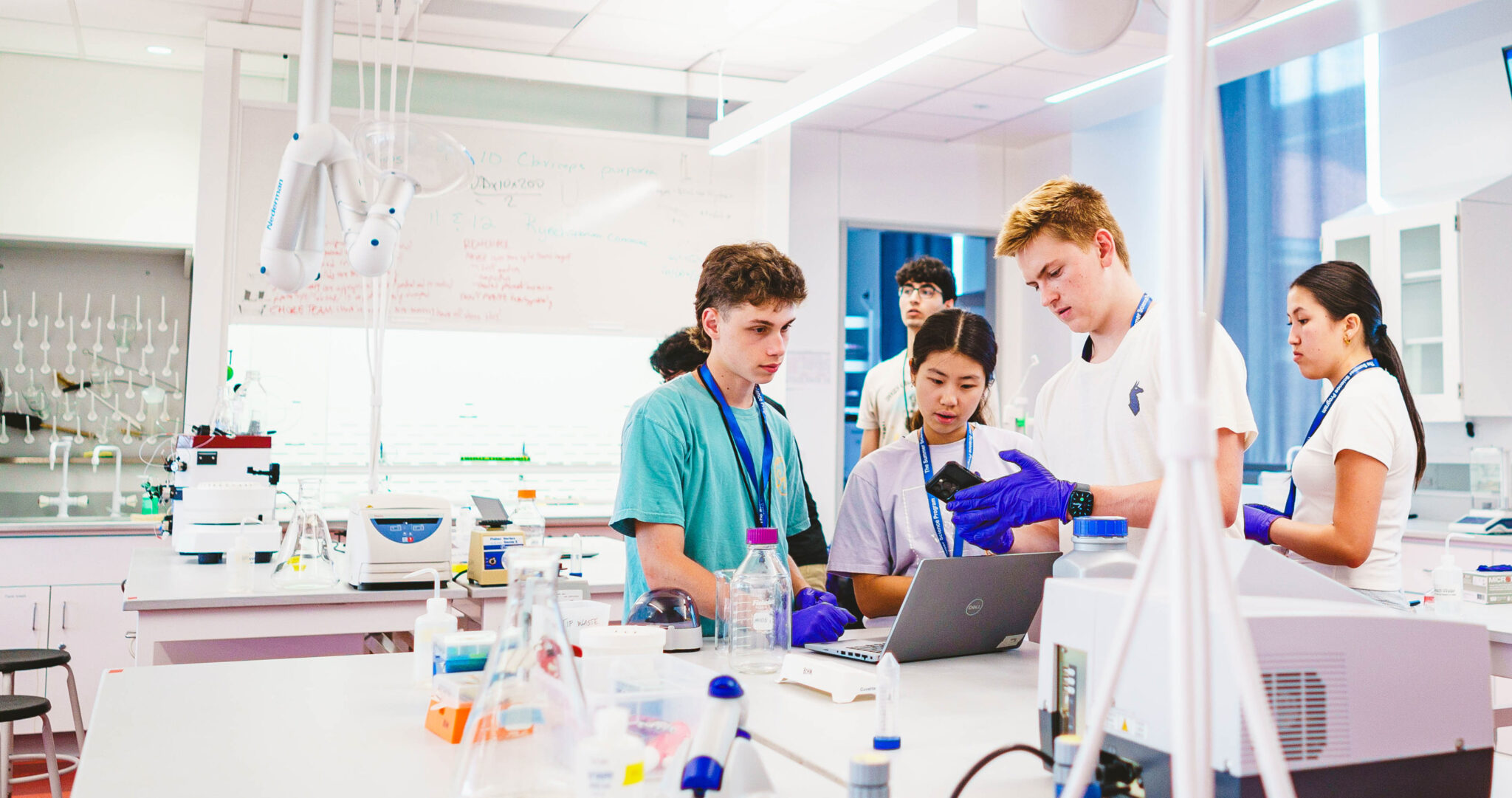 Five students in a laboratory collaborate on an experiment using a laptop, surrounded by scientific equipment and glassware. With financial aid supporting their ambitions, they dive into groundbreaking research with focus and determination.