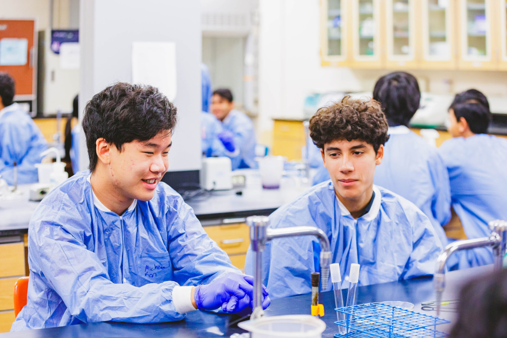 Two students in blue lab coats sit at a lab table with test tubes and equipment, engaged in discussion. Others work in the background.