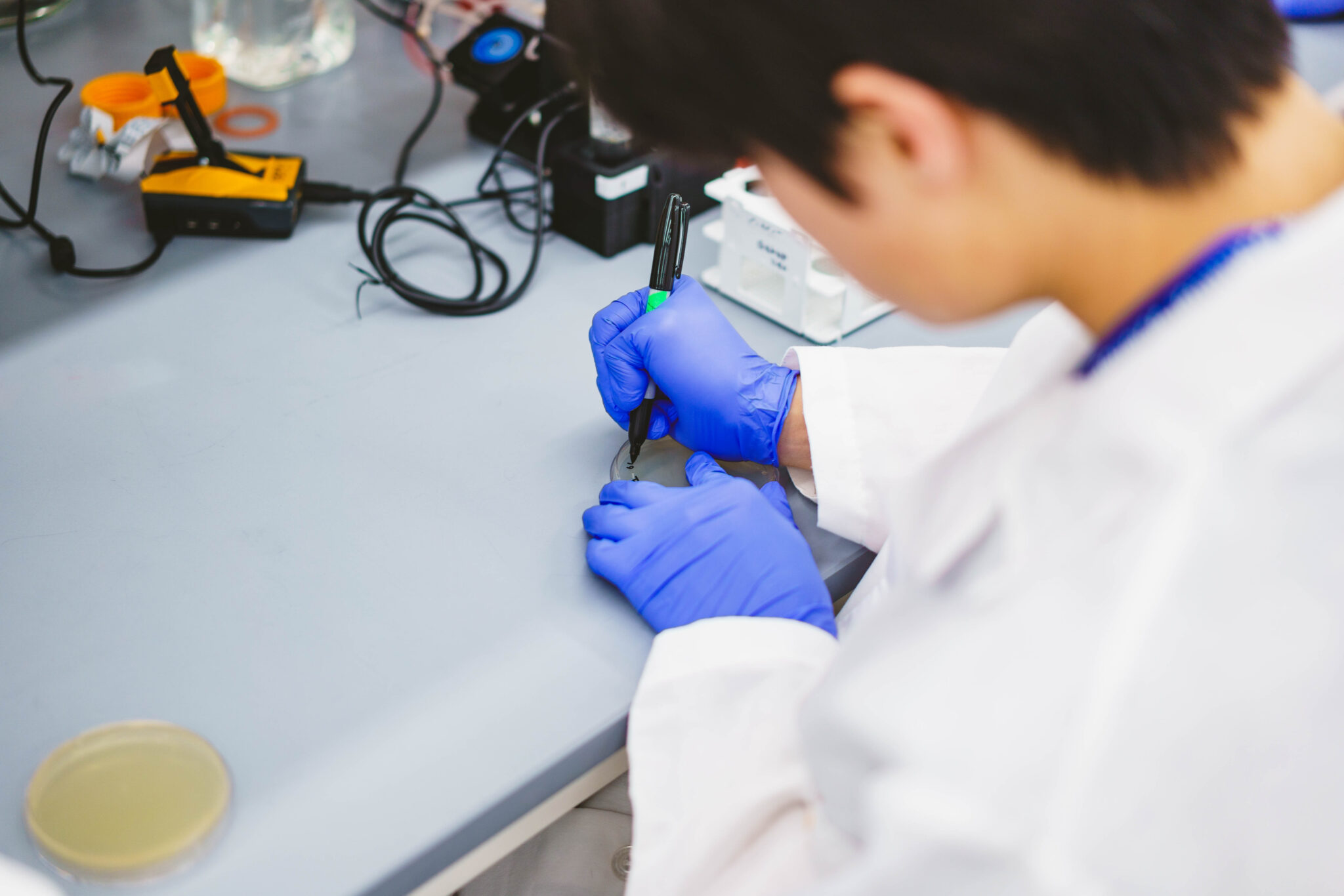 A person in a lab coat and blue gloves writes on a small object at a lab bench with various scientific equipment nearby.