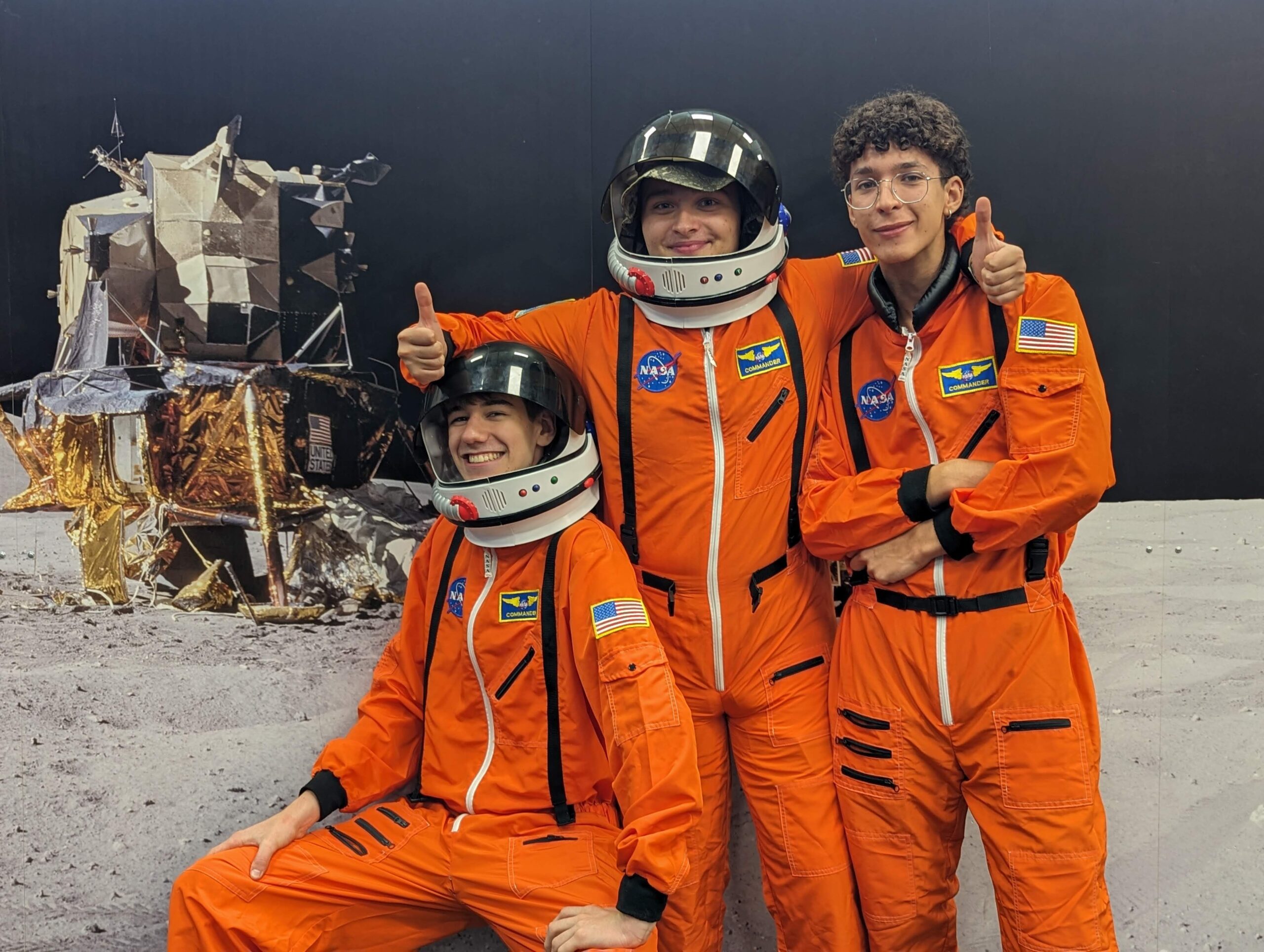 Three people in orange astronaut suits stand smiling in front of a lunar module backdrop, capturing the adventurous spirit of campus life. One gives a thumbs up, embodying the excitement and camaraderie found in college adventures.