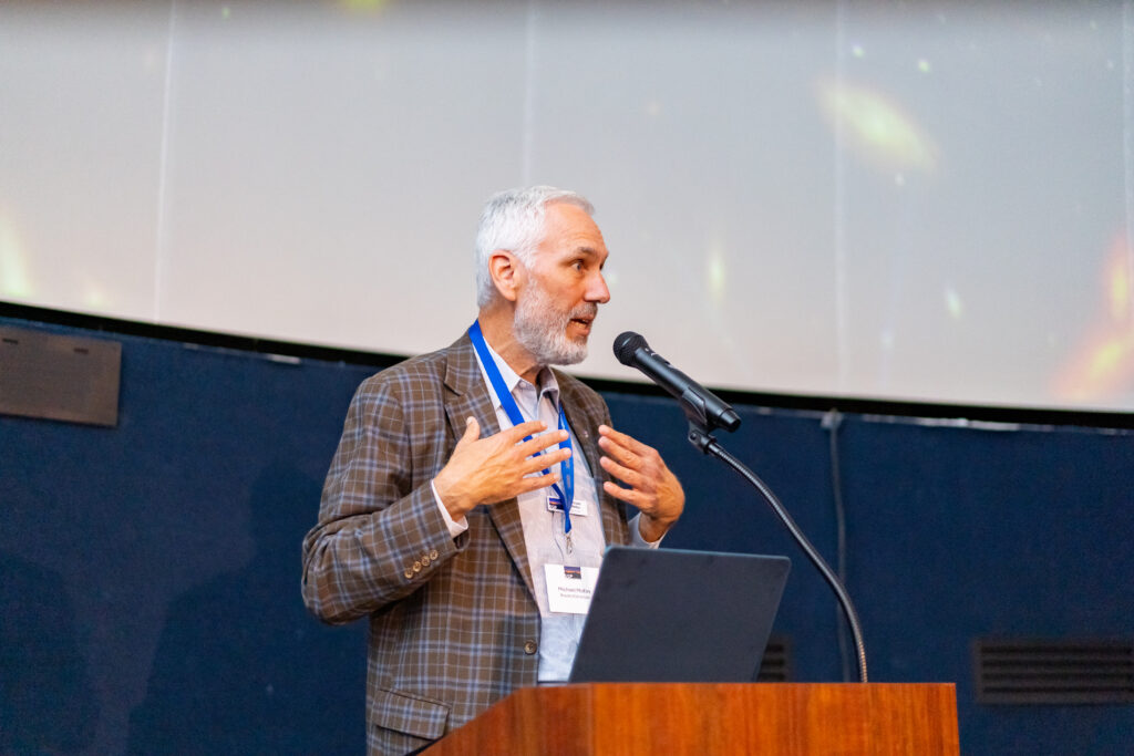 A man with a beard speaks into a microphone at a podium, gesturing with his hands. He wears a plaid blazer and name tag, with a projection screen behind him.
