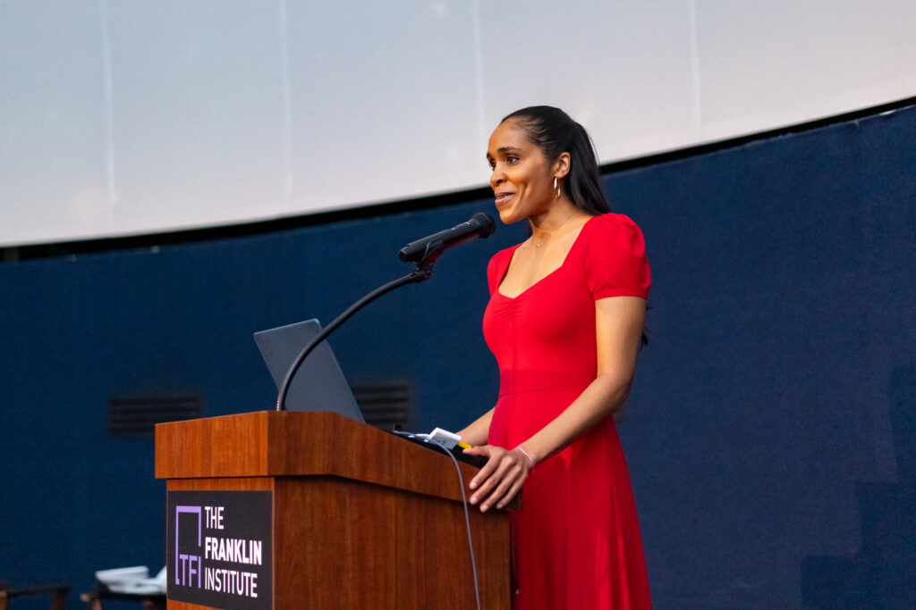 A person in a red dress speaks at a podium with a microphone, standing in front of a blue and white background.