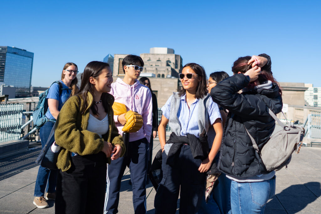 A group of people stand and converse on a rooftop with city buildings in the background.