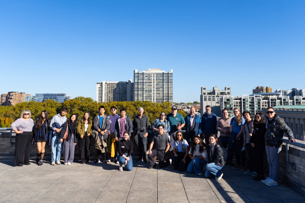 A group of people posing on a rooftop with city buildings and trees in the background under a clear blue sky.