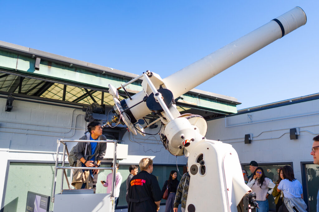 Person observing through a large telescope at an outdoor facility, while others watch nearby under a clear blue sky.