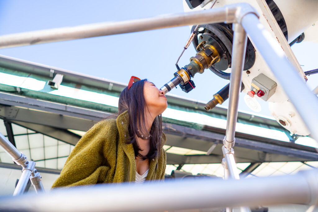 Person looking through a large telescope outdoors, surrounded by metal railings and a clear sky in the background.