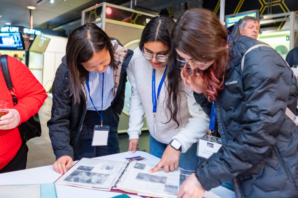 Three people examine a scrapbook with photographs and documents. They wear name badges and winter jackets.