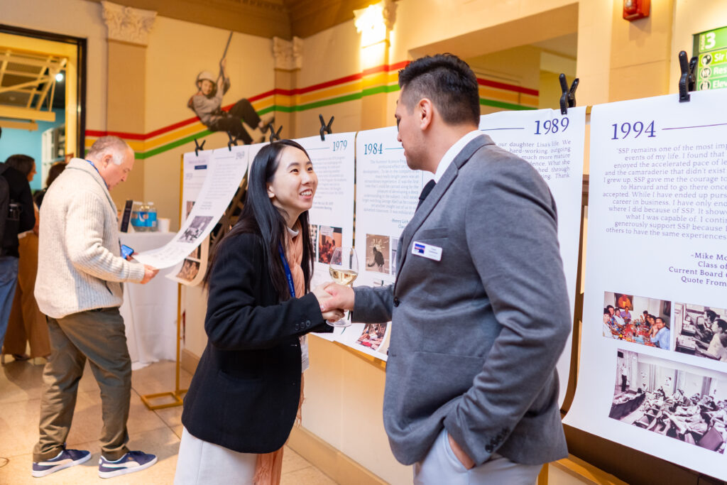 Two people shaking hands at an exhibition with informational boards. A man in the background reads a display.