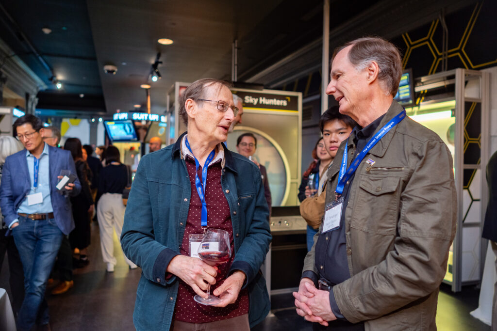 Two men with conference badges talk while holding drinks in a crowded room. People and exhibition booths are visible in the background.