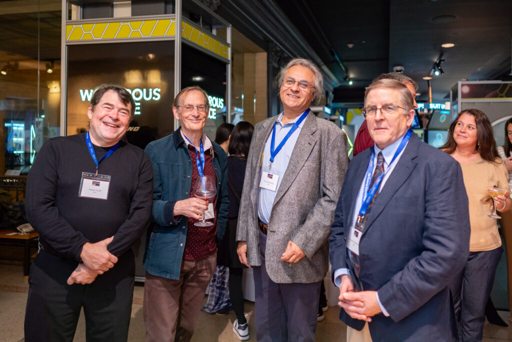 Four men stand together at an event, wearing name badges and smiling. Two hold drinks. People are gathered in the background.