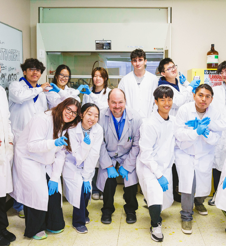 A group of people in lab coats and gloves pose in a laboratory setting, smiling at the camera.