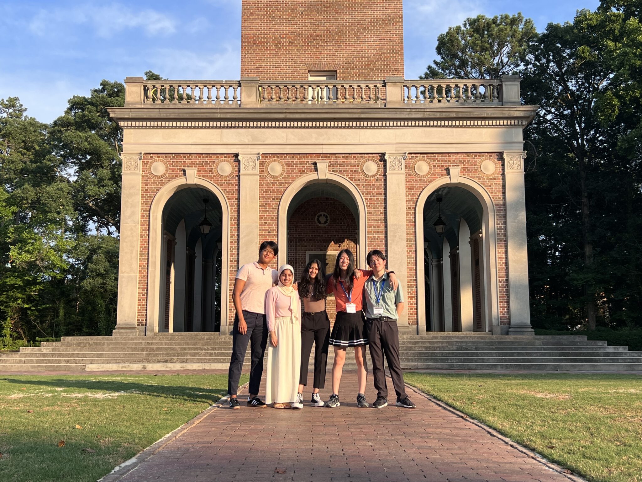 Five people standing in front of a brick building with arches, surrounded by trees, under a clear blue sky.