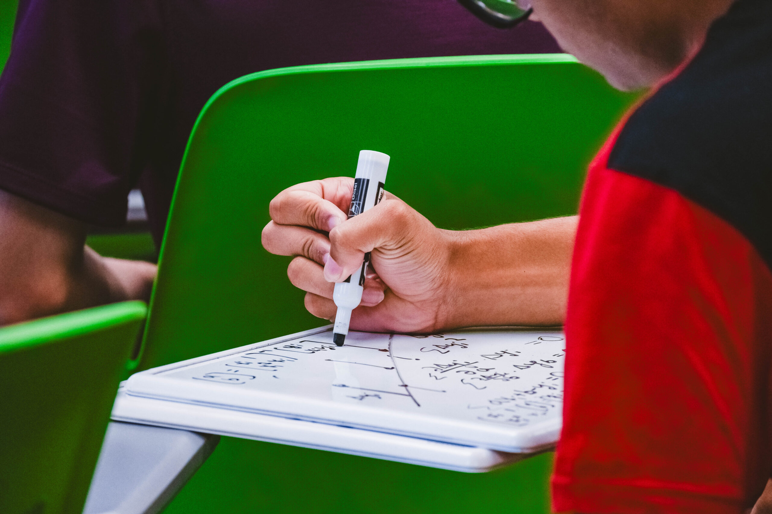 A person writes on a small whiteboard with a black marker, surrounded by green chairs.