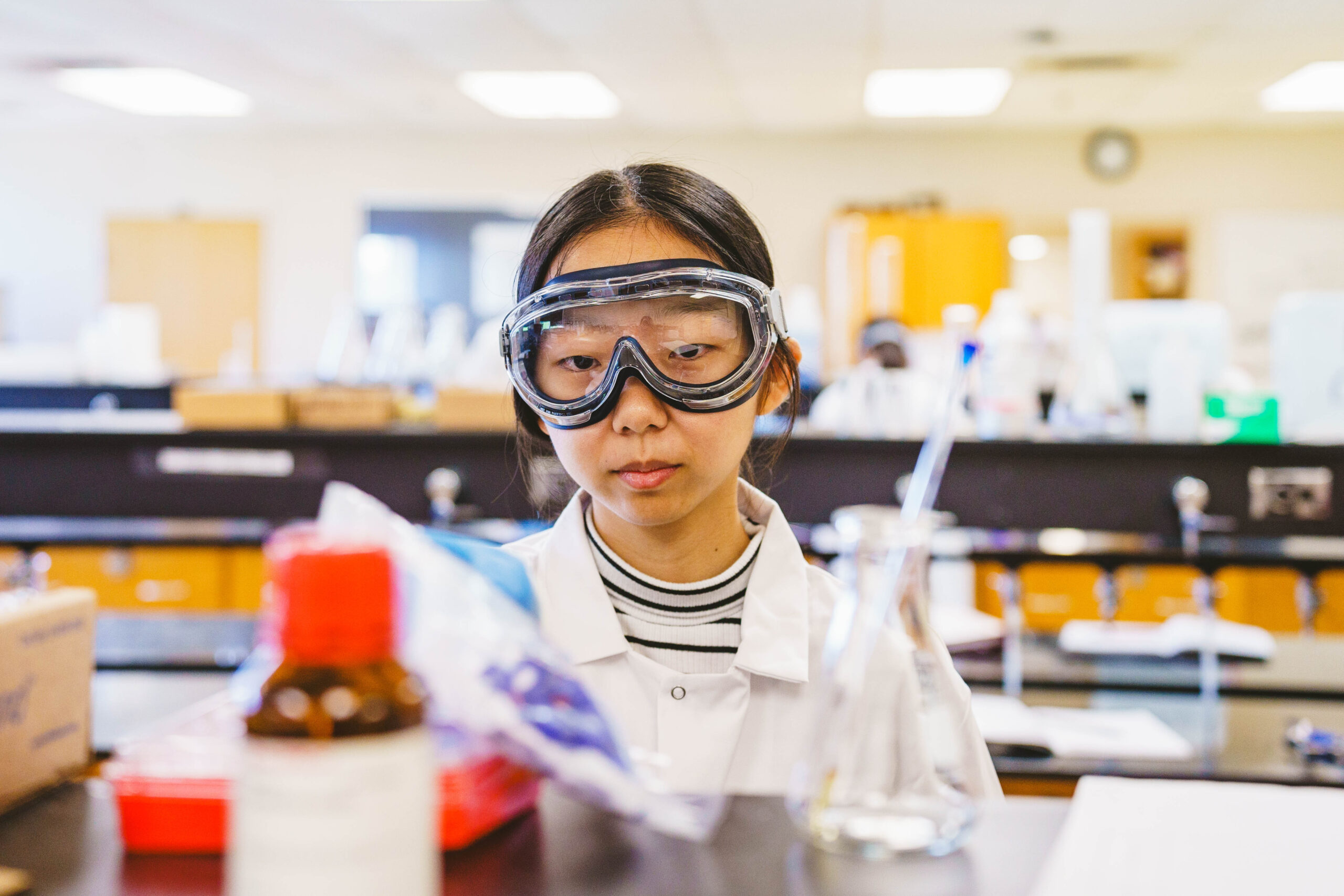 A person wearing protective goggles and a lab coat conducts an experiment in a laboratory, surrounded by equipment and bottles.
