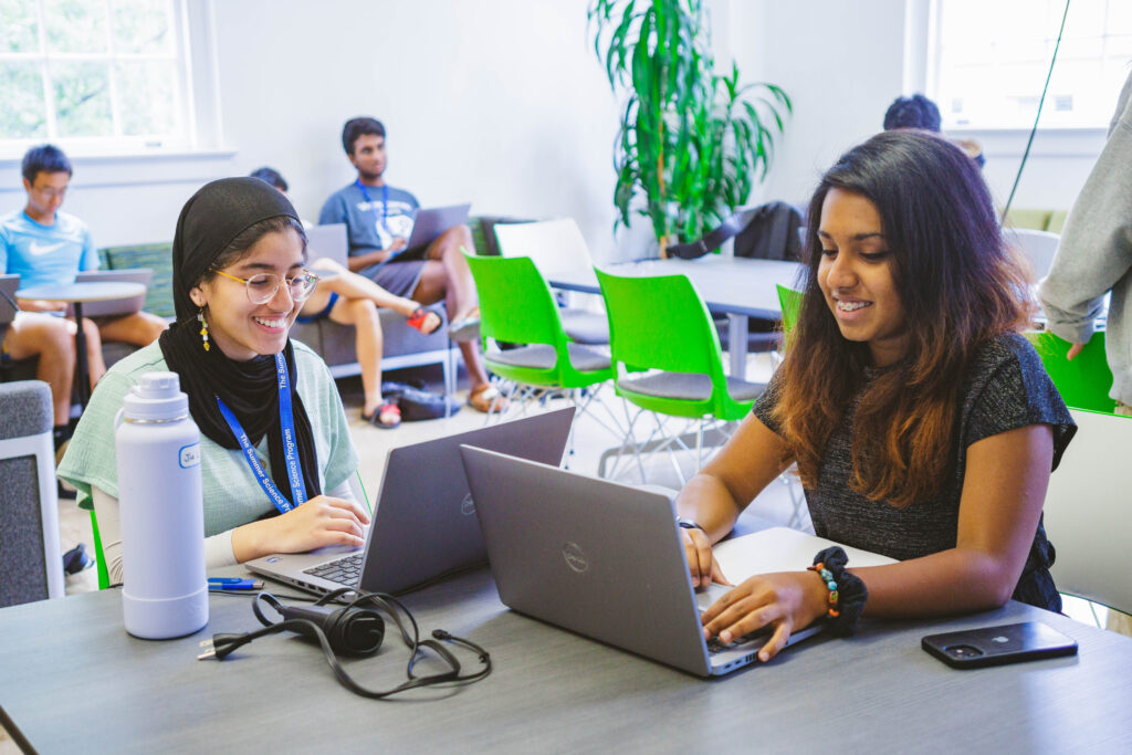 Two people sit at a table using laptops in a room with green chairs and other people working in the background. A large plant is visible near the window.