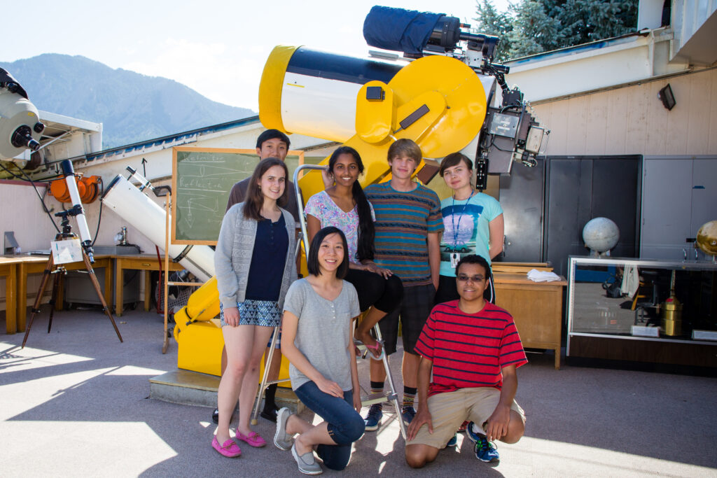 Group of seven people posing in front of a large yellow telescope in an outdoor observatory setting.