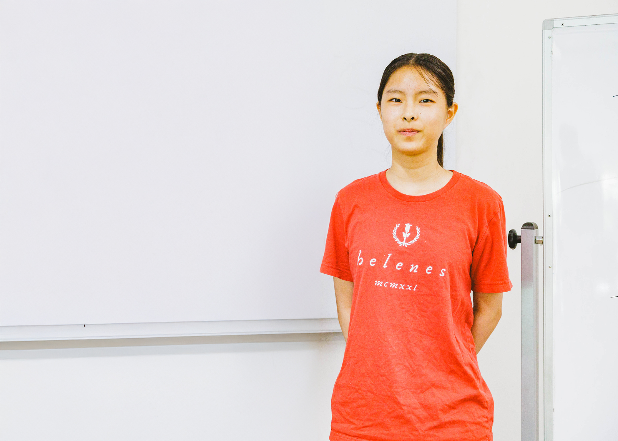 A person stands in a room wearing a red shirt with text, posing in front of a whiteboard.