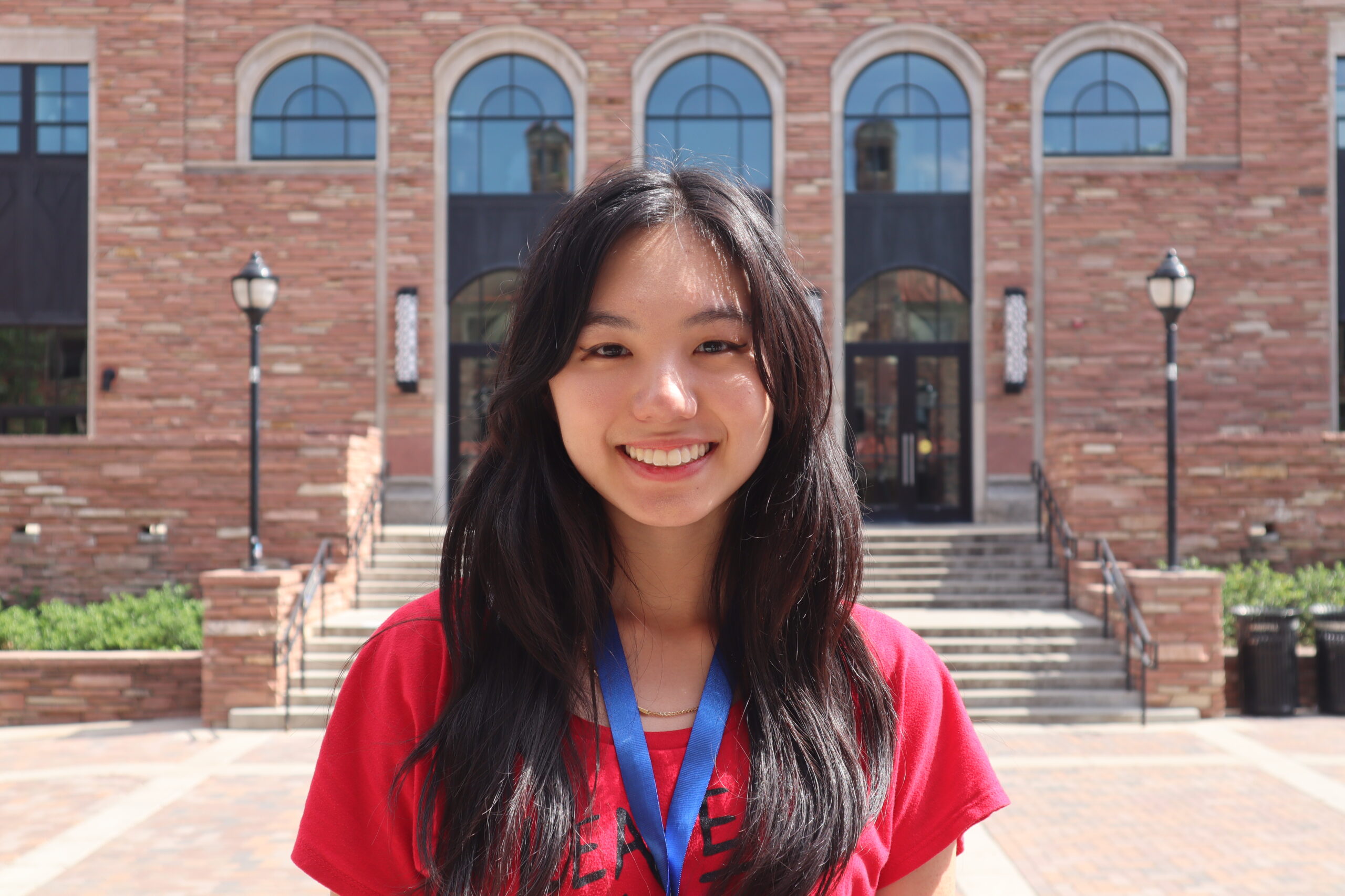 A person with long dark hair smiles in front of a brick building with large windows and multiple archways. They are wearing a red shirt and a lanyard.