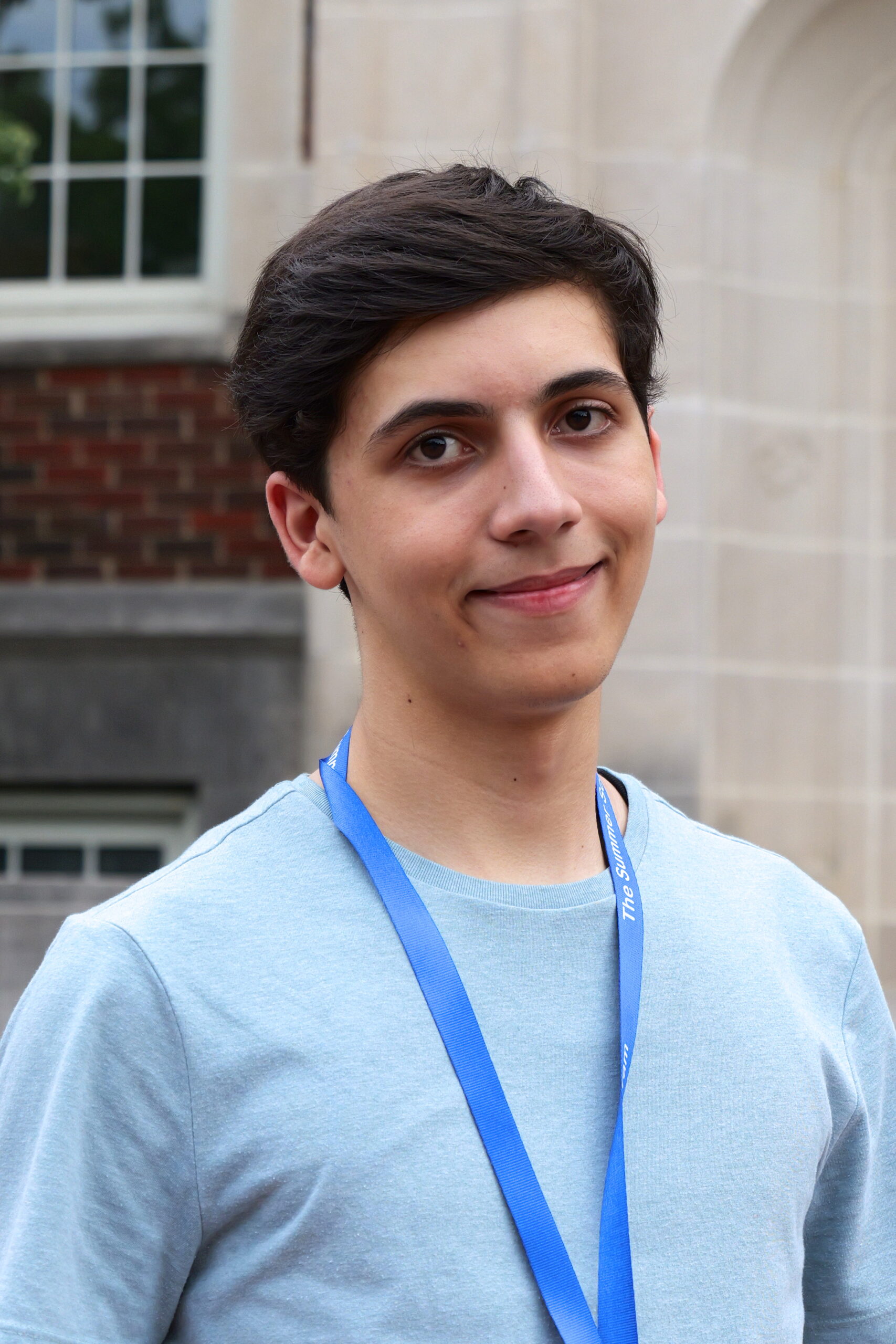 Young person wearing a light gray shirt and blue lanyard, standing outside in front of a building with brick and stone walls.