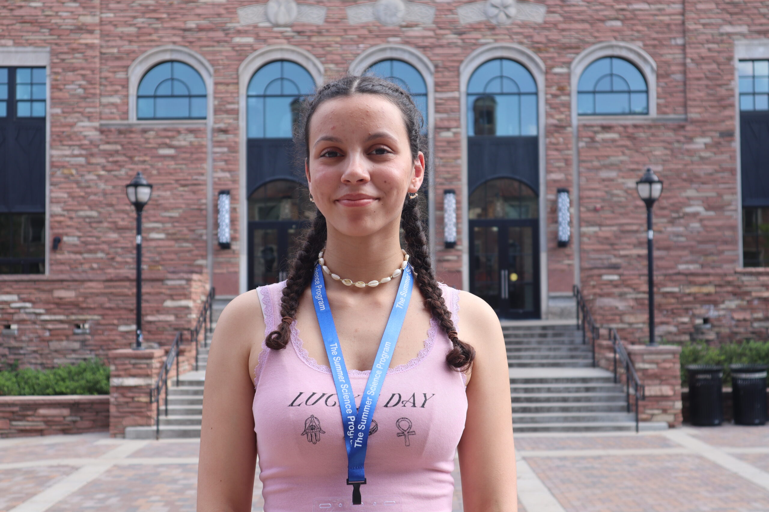 A person with braided hair stands outside a brick building with arched windows, wearing a pink tank top and a blue lanyard.