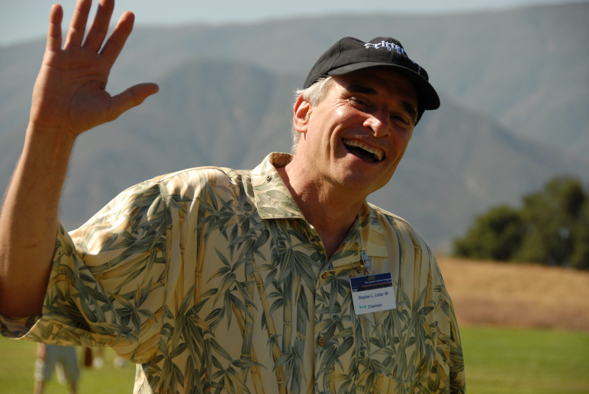 A person wearing a black cap and patterned shirt smiles and waves outdoors with mountains in the background.