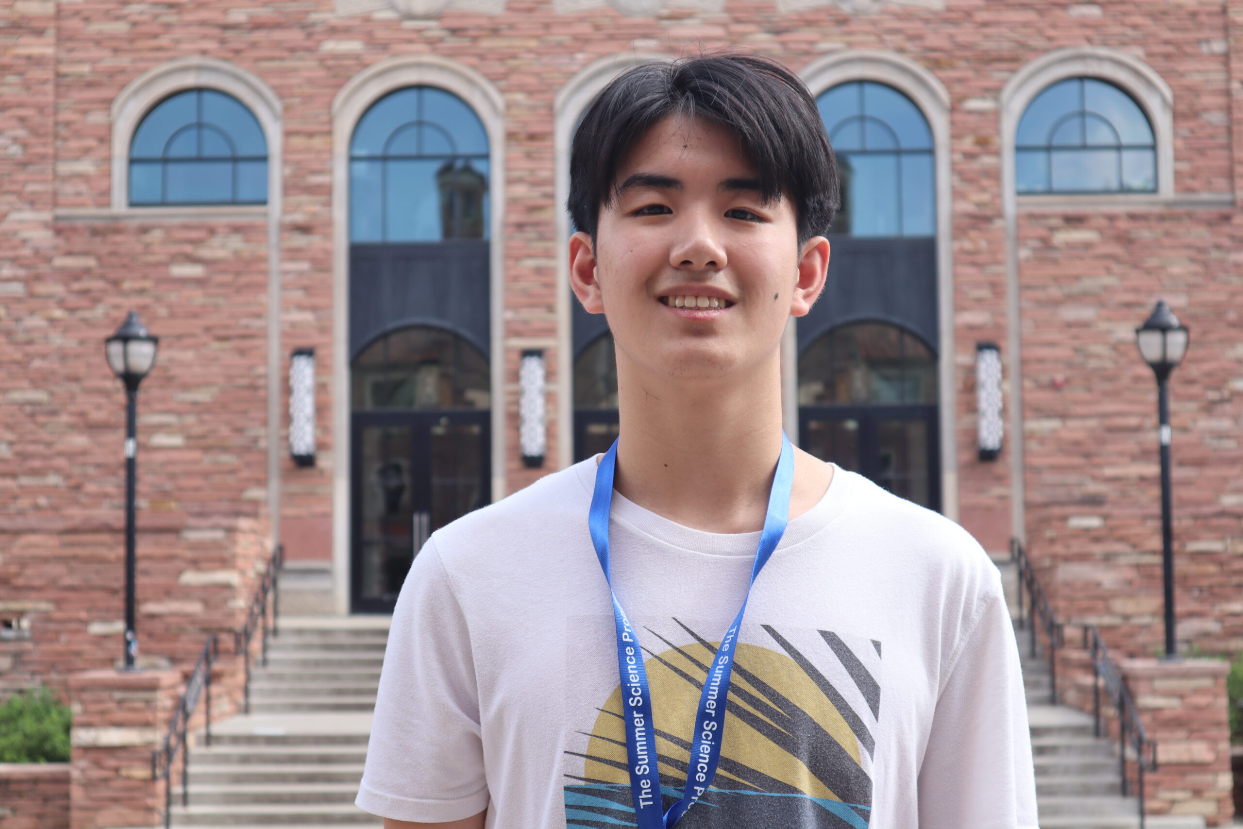 A person wearing a white t-shirt and blue lanyard stands smiling in front of a brick building with arched windows.