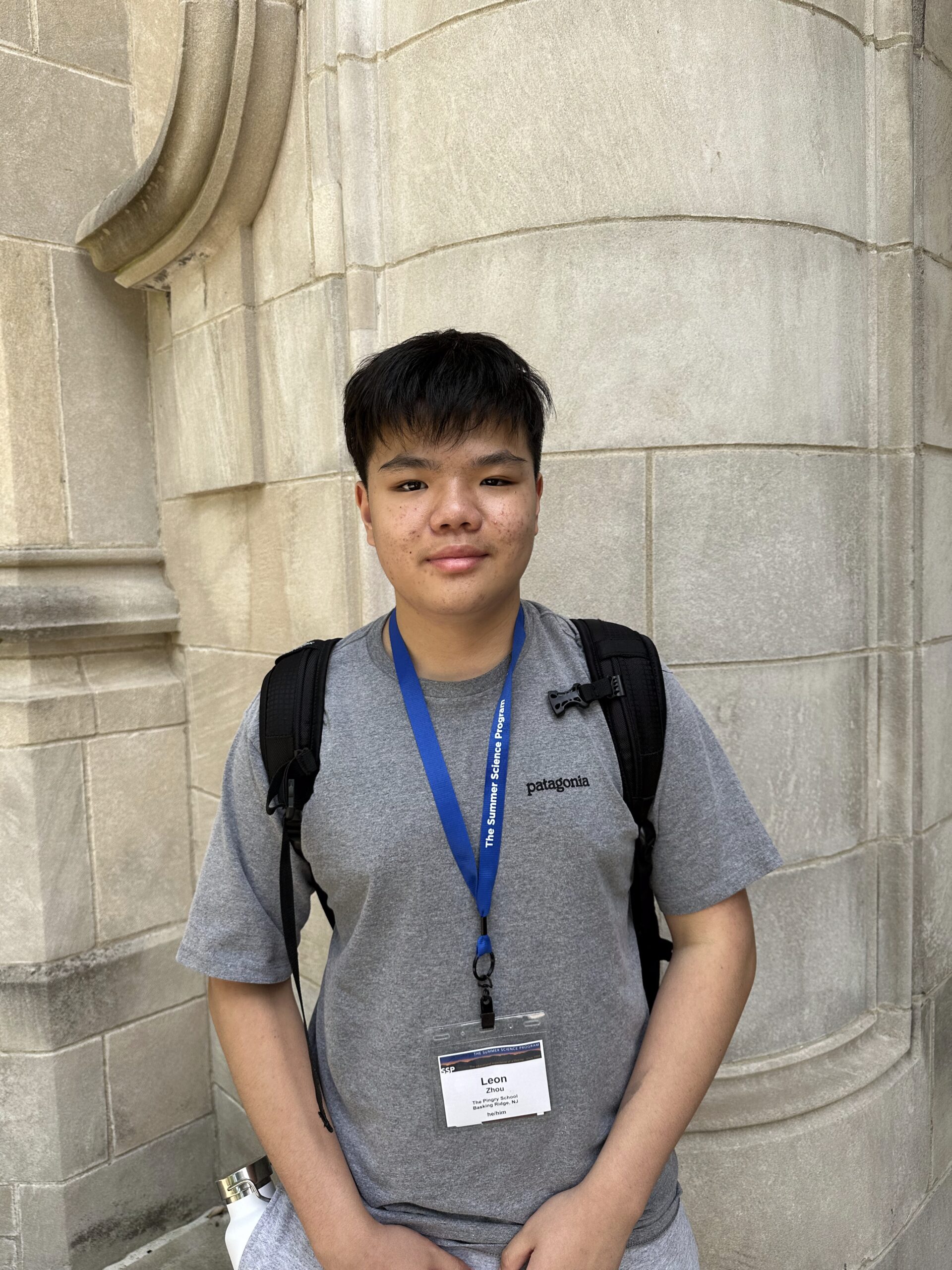 A person wearing a gray T-shirt and a blue lanyard with a name tag stands in front of a stone wall, carrying a black backpack.