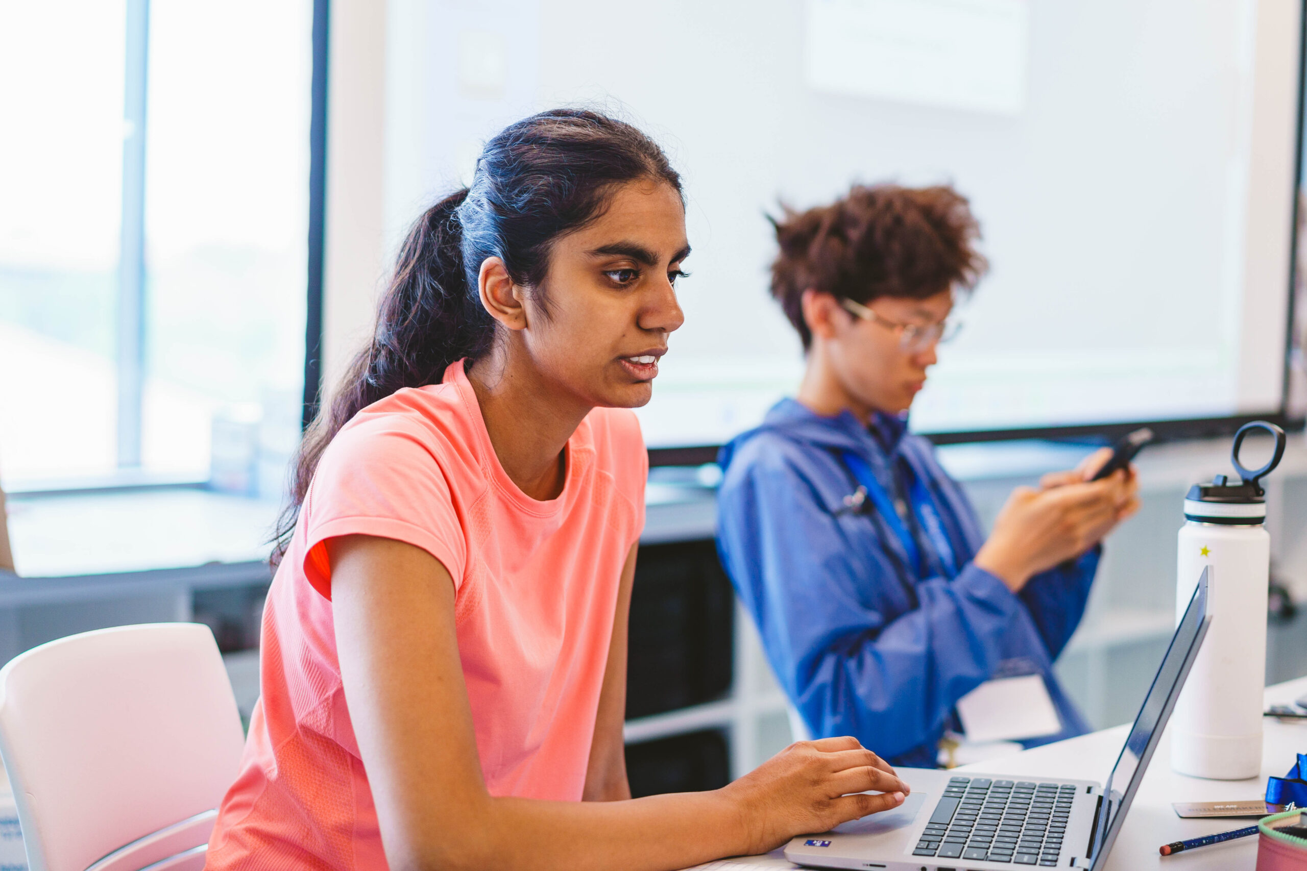 A person in a pink shirt works on a laptop, while another in a blue jacket uses a phone. They are indoors with a bright window in the background.