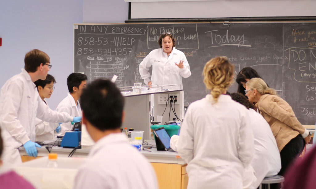 A group of people in lab coats are engaged in a science class. A person at the front gestures while others work at desks. A chalkboard with notes is visible in the background.