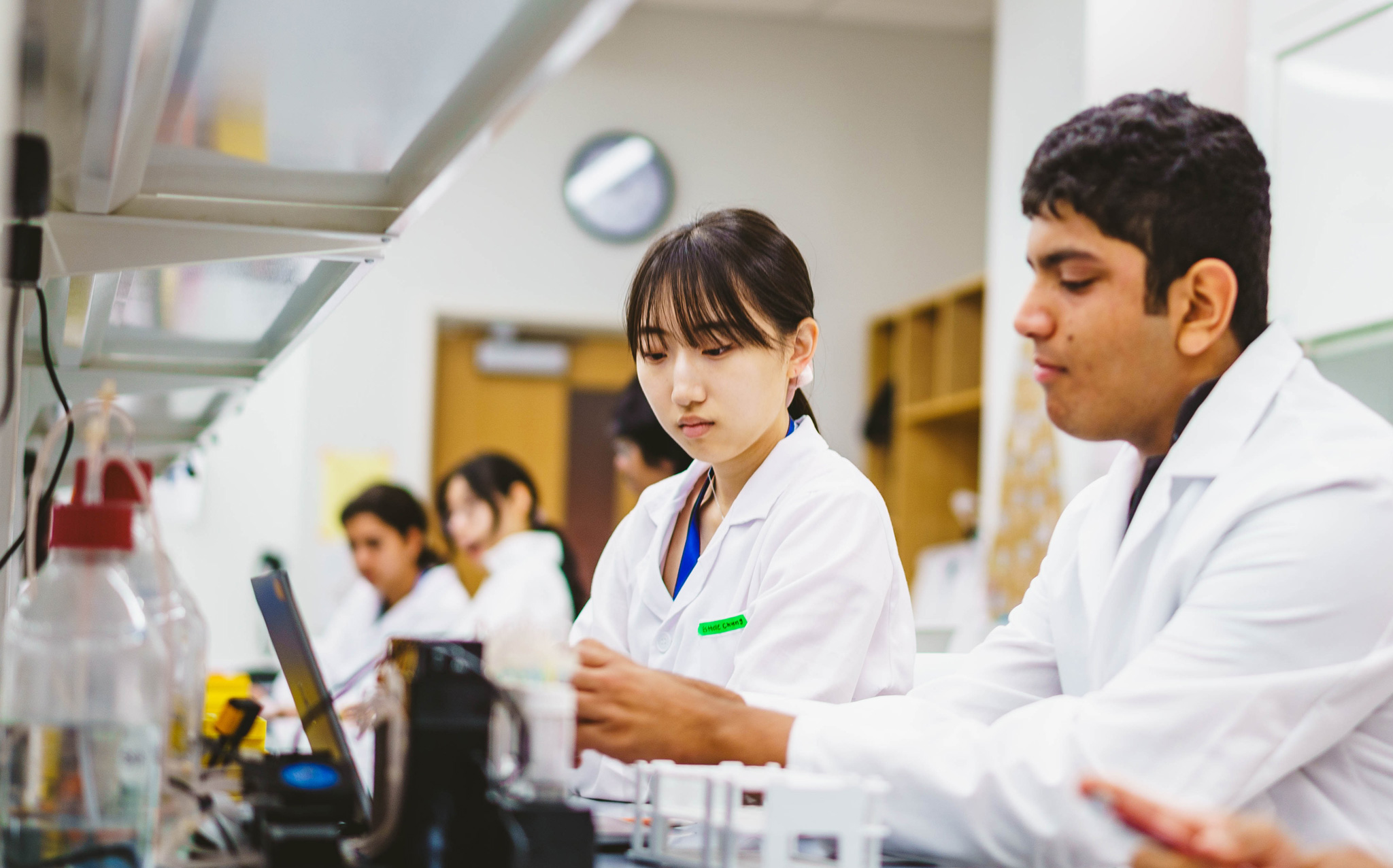 Scientists in white lab coats work at a laboratory bench with equipment and computers, focusing on their tasks.