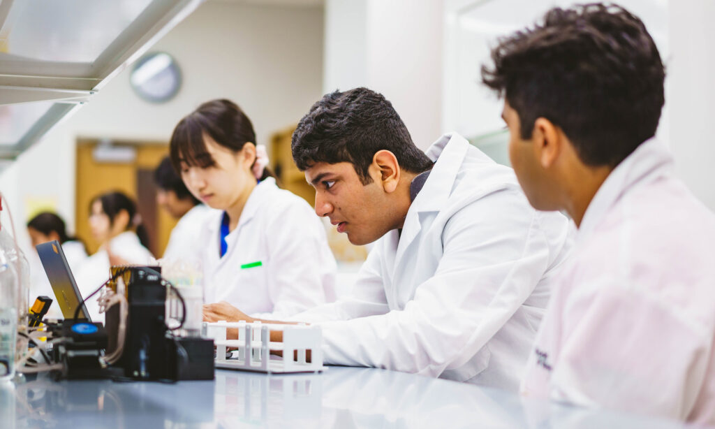Three scientists in white lab coats are working in a laboratory. One is focused on a laptop, while others are engaged in their tasks. Laboratory equipment is visible on the table.