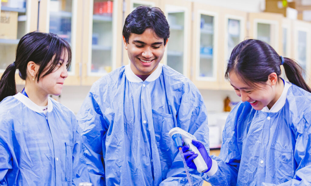 Three people in blue lab coats work together in a laboratory, using a pipette while smiling and discussing their task.