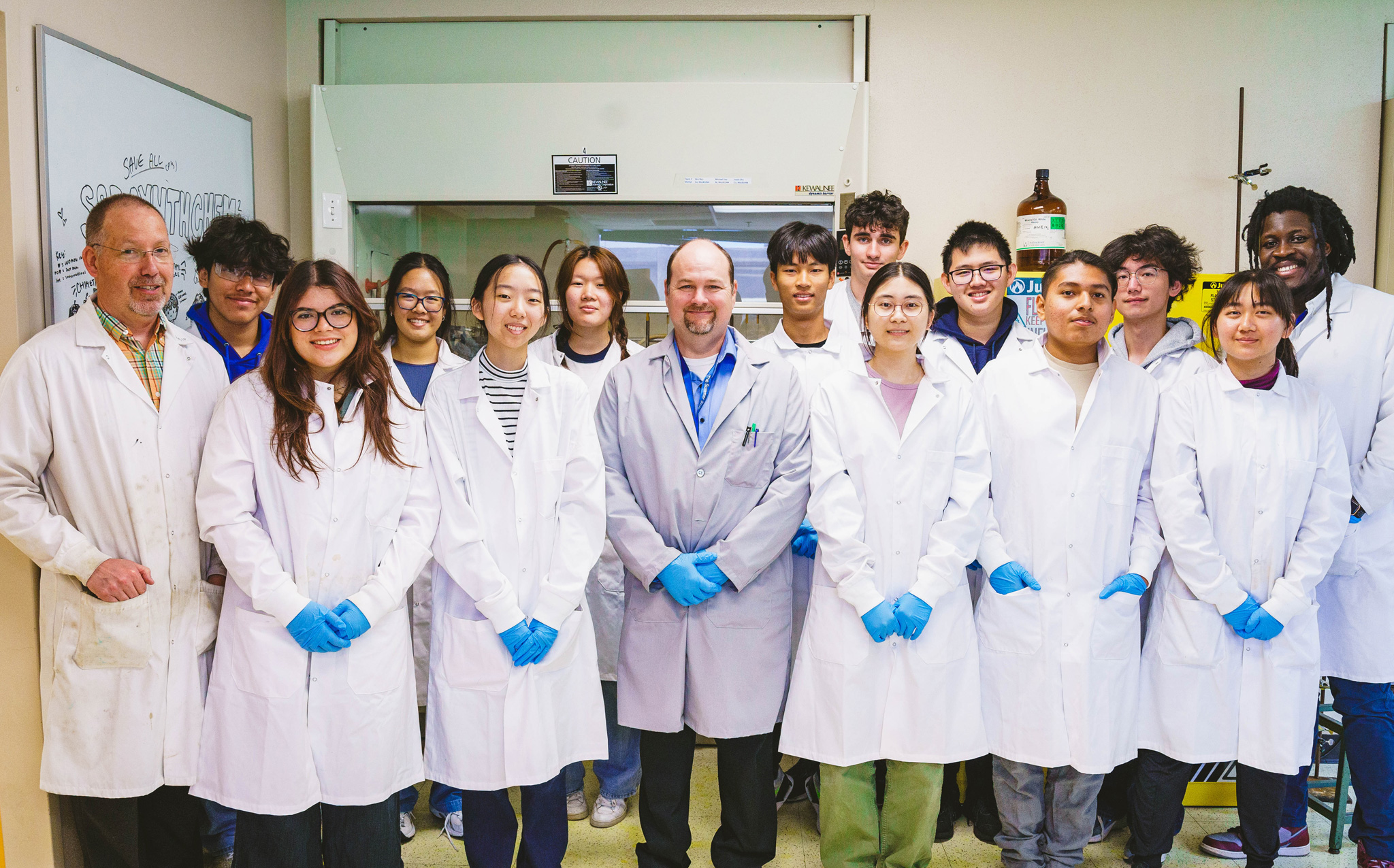 A group of people wearing white lab coats and blue gloves pose for a photo in a laboratory setting.