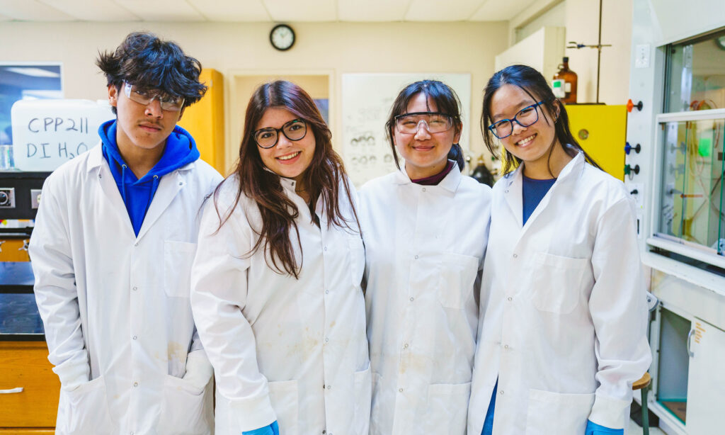 Four people wearing lab coats and safety goggles stand together in a laboratory.