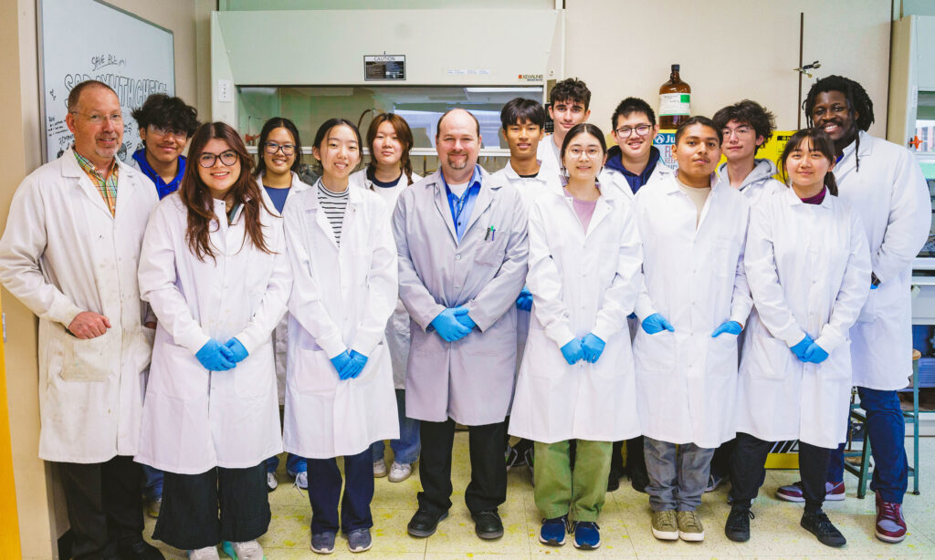 A group of 15 people in lab coats and blue gloves stand together in a laboratory setting, posing for a group photo.