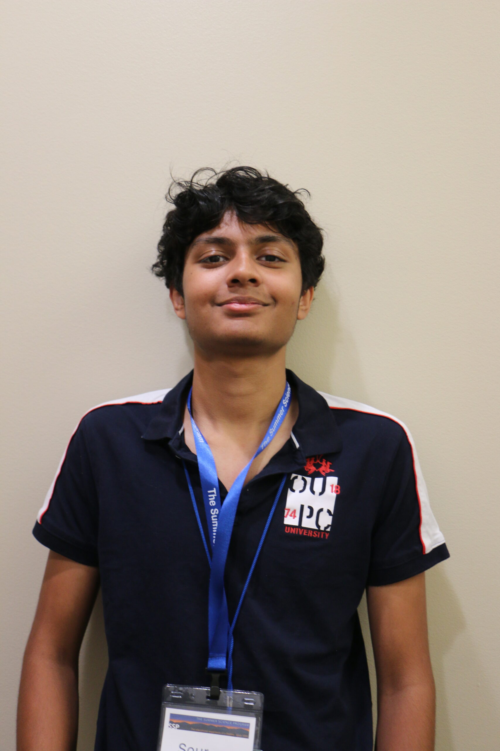 A person stands in front of a beige wall, wearing a navy polo shirt with a university logo and a blue lanyard with a name badge.