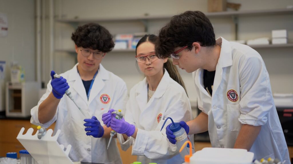 Three individuals in lab coats and gloves are working with pipettes in a laboratory setting.
