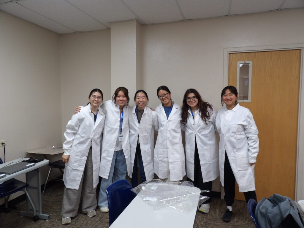 Six people wearing white lab coats stand together in a room, smiling at the camera.