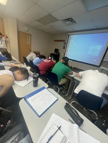 People in a classroom appear to be napping at desks, facing a projected screen with a Windows desktop.