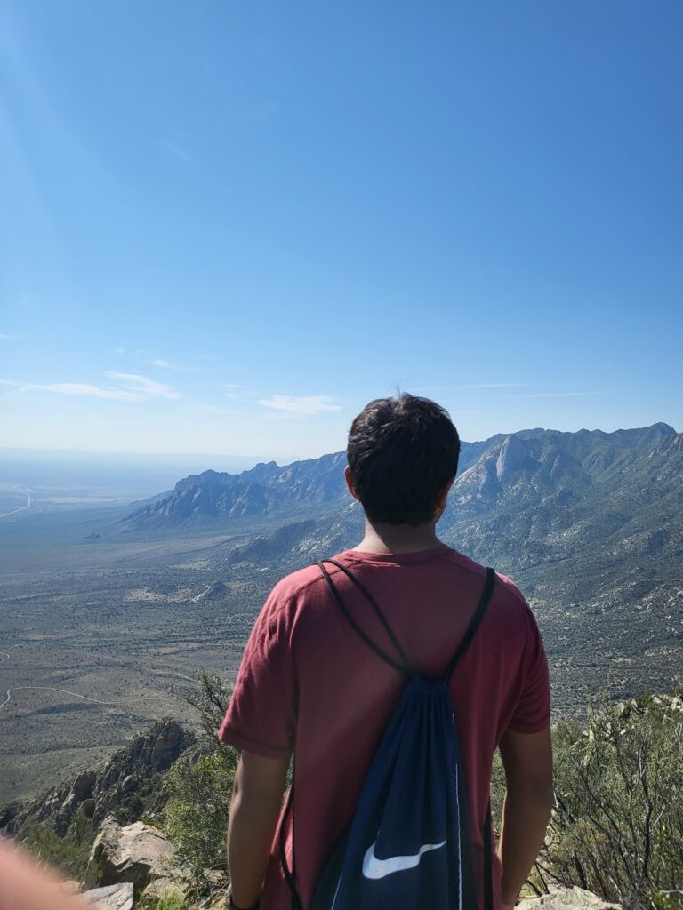 Person in a red shirt with a dark blue drawstring bag stands on a mountain overlook, gazing at a vast landscape of mountains and plains under a clear blue sky.
