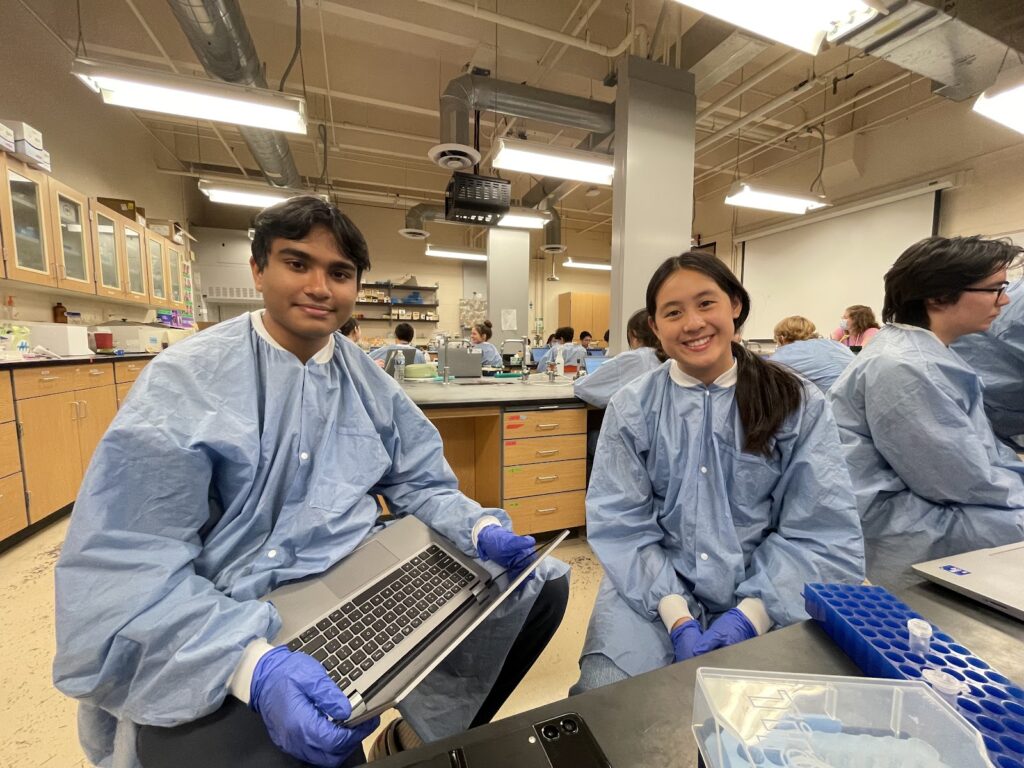 Two individuals in blue lab coats and gloves sit at a lab desk with a laptop and lab equipment, smiling at the camera.