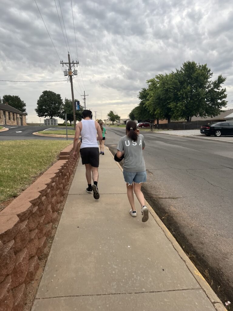 Two people walking on a sidewalk under a cloudy sky. The person on the left wears a white tank top and shorts, while the person on the right wears a gray