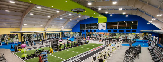 A spacious gym with equipment including weights, cardio machines, and exercise balls, under a high ceiling with bright lighting.