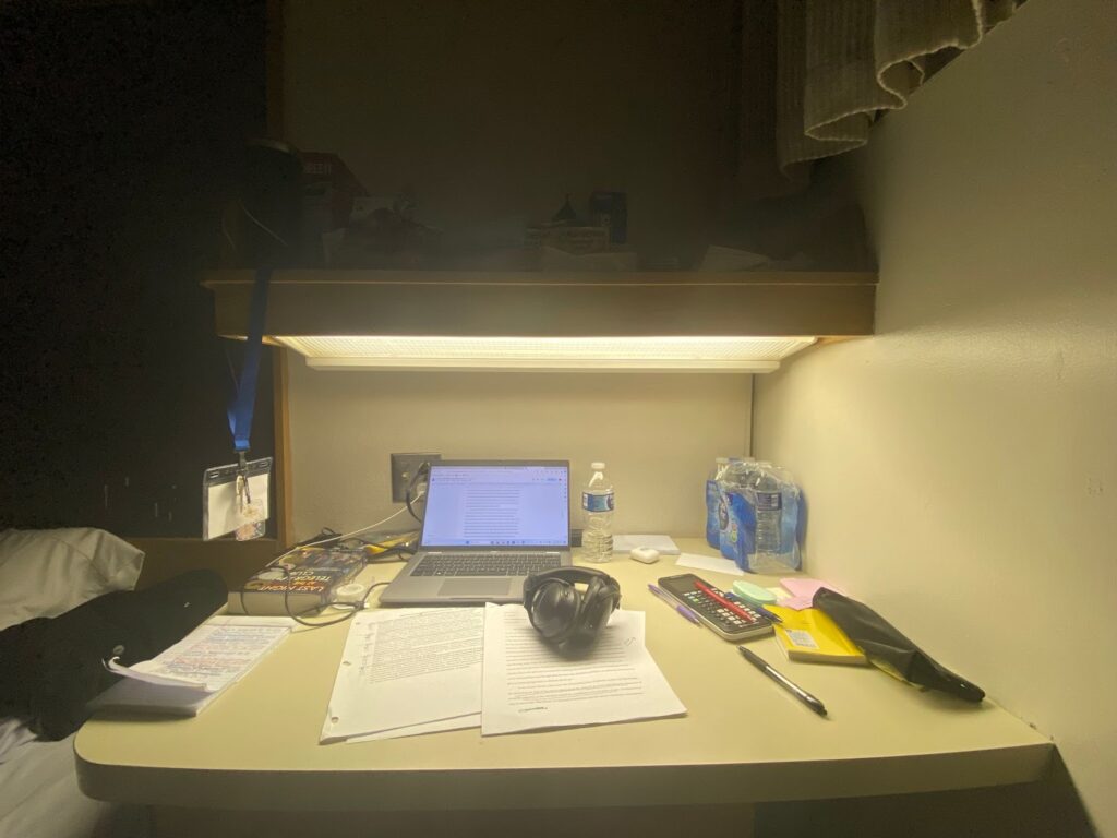 A study desk with a laptop, papers, a calculator, headphones, a water bottle, books, and a shelf above.