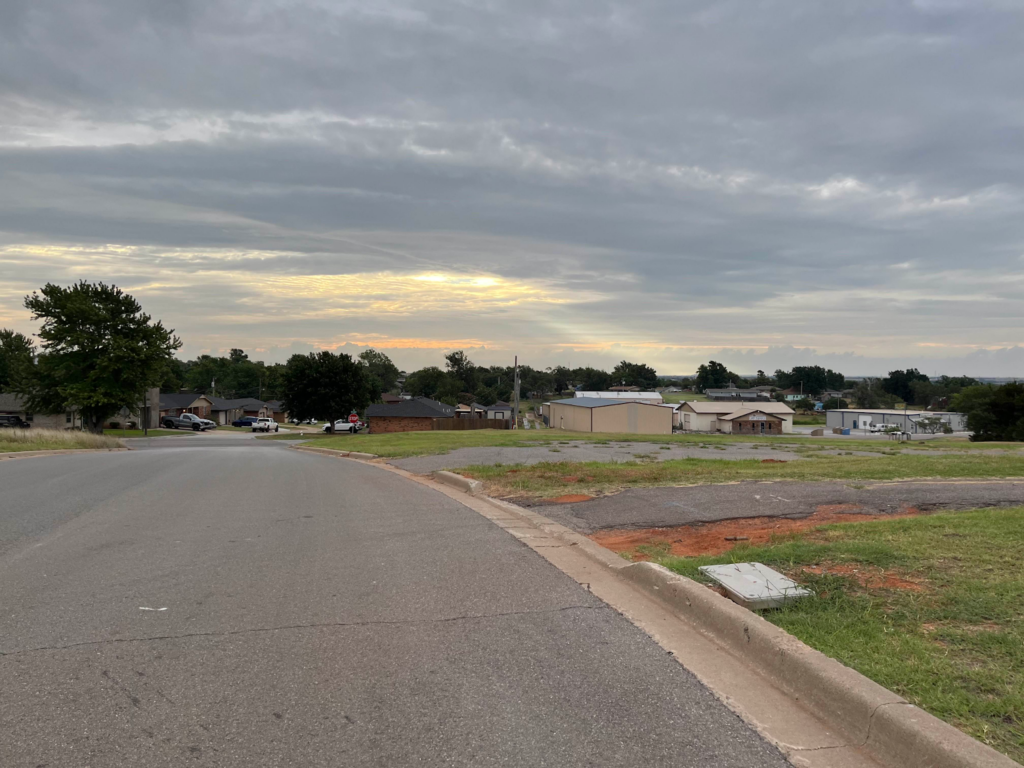 Suburban street under a cloudy sky with a setting sun, featuring houses, trees, and a patch of grass on the roadside.