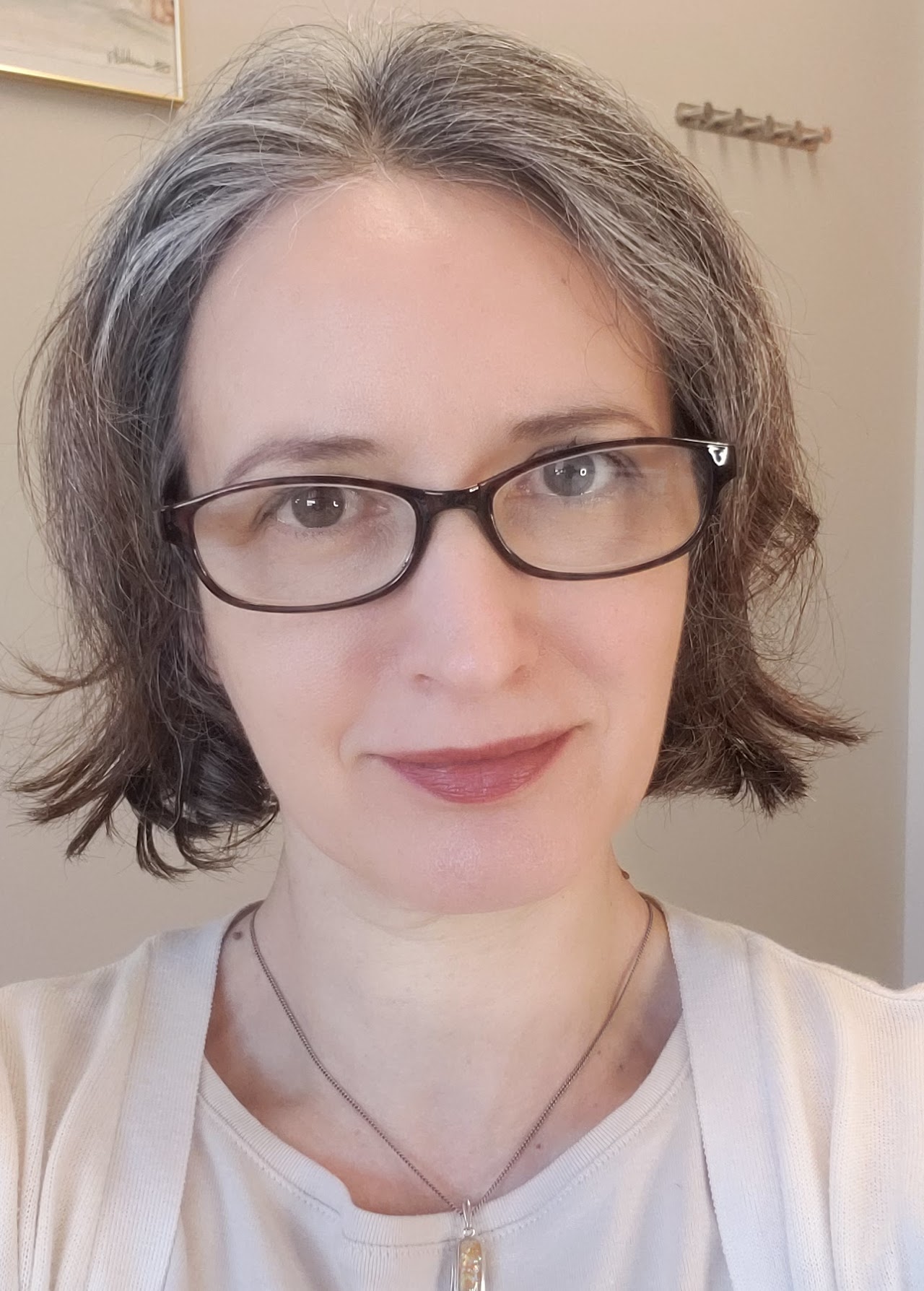 A person with short, wavy gray hair and glasses, wearing a light-colored top, is looking at the camera with a neutral expression.