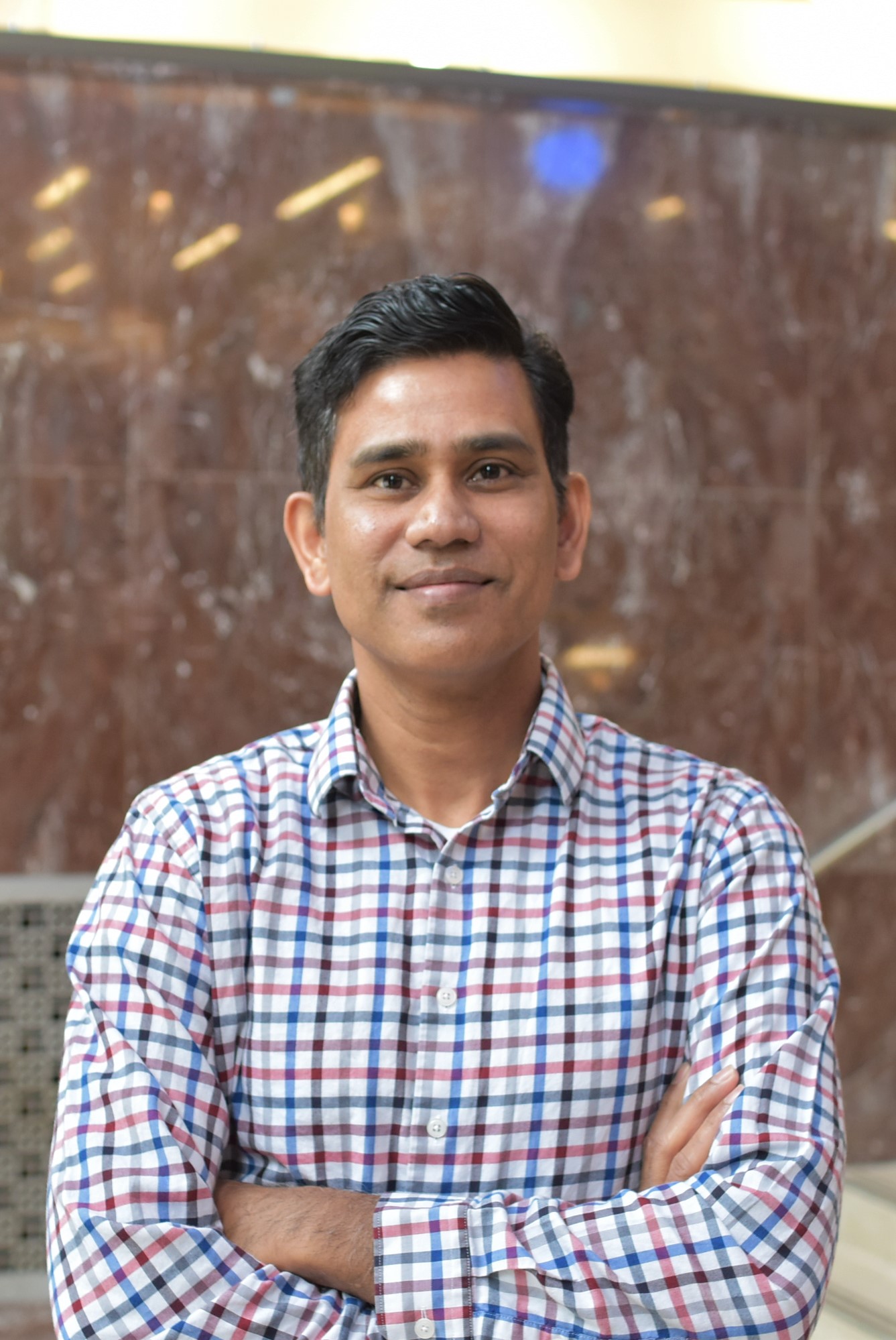 A person with short dark hair, wearing a plaid shirt, stands with arms crossed in front of a marble wall.