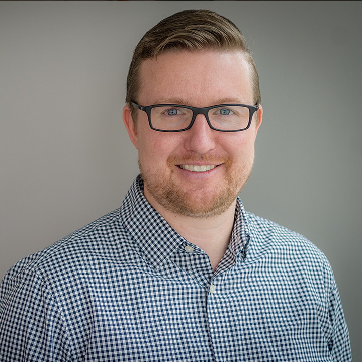 A man with glasses and a short beard is smiling, wearing a checkered shirt against a plain gray background.
