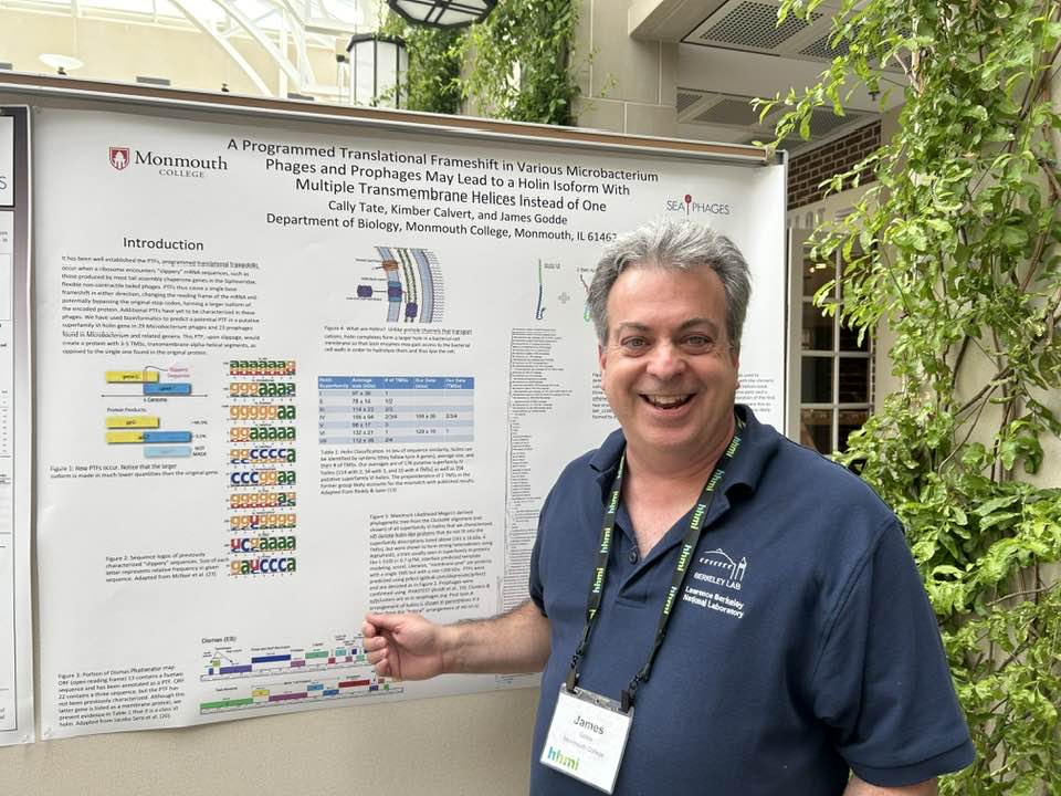 A person stands in front of a research poster, pointing at a section. The poster is displayed at an indoor venue.