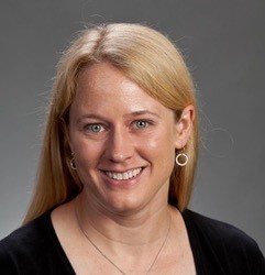 A person with long blond hair, smiling, wearing a black top and hoop earrings, is posed against a plain gray background.